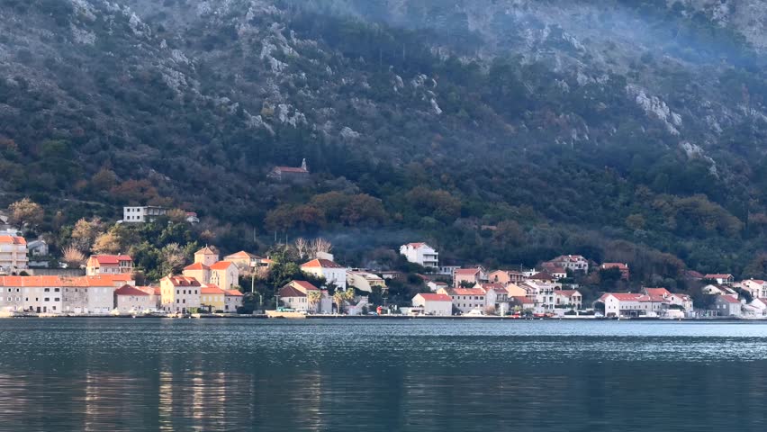 Calm seaside view of a small coastal village with traditional houses along the shoreline, reflected
in still water. Forested mountains rise behind the town, creating a quiet, atmospheric scene o f
slow life, travel, and Mediterranean coastal nature.
