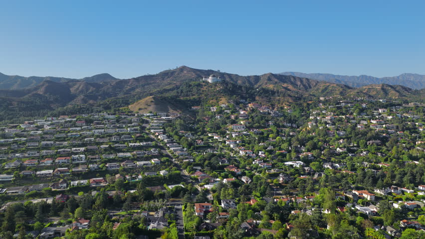 Aerial drone panorama of Los Angeles California showing dense urban neighborhoods blending into green hills and distant mountains under bright daylight conditions. Griffith observatory on a background