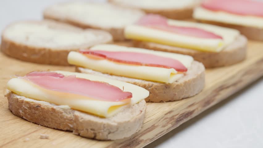 Hands closing prepared sandwiches with cheese and ham on a wooden board, food preparation process.