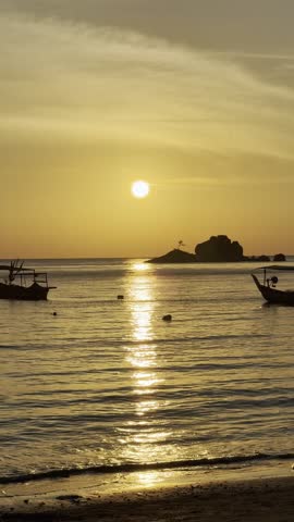 Golden sunset over a calm ocean with silhouettes of traditional fishing boats and a small island. Peaceful waves washing on the sandy beach during golden hour. Vertical video.