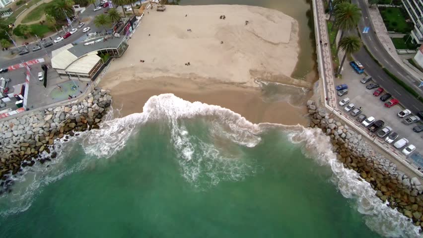 drone view of a coastal landscape where ocean waves meet a sandy beach between two rocky breakwaters. The scene shows turquoise water, white sea foam, shoreline textures, urban promenade, and a small river outlet flowing into the sea. Static overhead composition highlighting natural patterns, coastal erosion control, and the interaction between city infrastructure and the ocean.