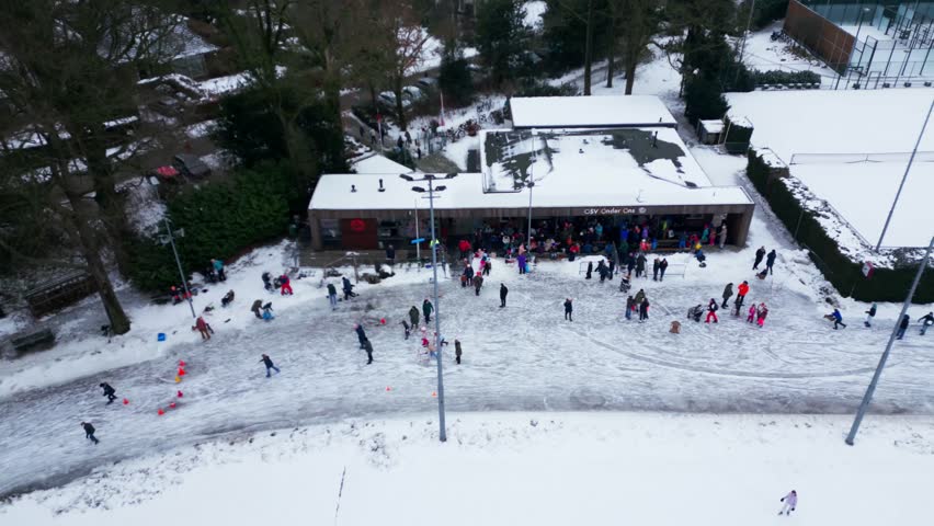 Aerial drone shot of the ice skating clubhouse and adjacent snowy courts in Oosterbeek. People gathering near the building for winter sports. Snow covered park landscape in the Netherlands.
