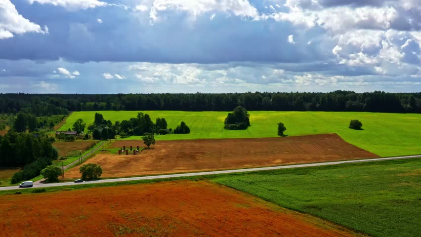 Aerial view of green and brown farming fields, a country road, and forest under dynamic clouds