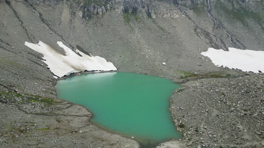 Cinematic drone circles a turquoise iceberg lake near Whistler, Canada, with snow patches, rocky gravel hills, steep mountain slopes, and small rivers flowing down, creating a cold, remote atmosphere