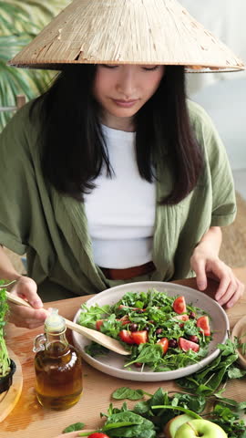 A woman wearing a traditional conical hat prepares a fresh salad with tomatoes and greens, with olive oil nearby