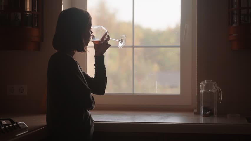 Elegant young woman drinking wine from a beautiful glass, standing in backlight on a modern kitchen. Female silhouette by the window creates a calm, cinematic evening lifestyle mood.