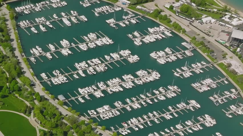 A marina in Chicago filled with boats docked in clear water. Nearby green areas and buildings add to the city scene during daytime.