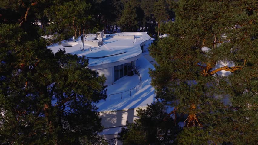 Aerial view shows a white terraced building in Jurmala, Latvia, amid tall pines. Snow covers the roof. Long blue shadows from winter sun add a cool quiet mood.