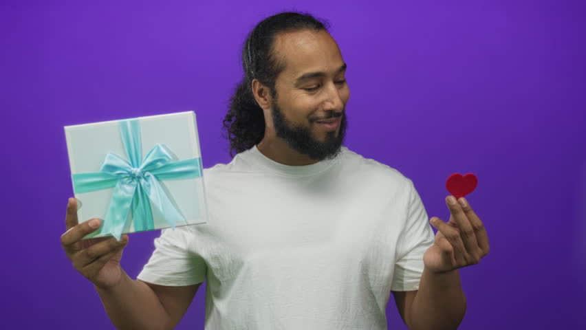 Man holding a gift box with blue ribbon and a small red heart between fingers, smiling in purple studio; love gratitude joy.