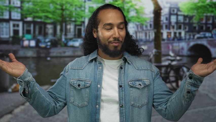 Man shrugging with open palms on an amsterdam canal street, smiling in denim jacket and white tee; serenity casual.