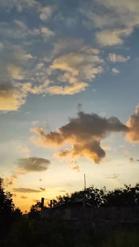 Golden sunset sky with scattered clouds glowing in warm light above dark silhouettes of trees and rooftops, creating a calm, atmospheric evening landscape