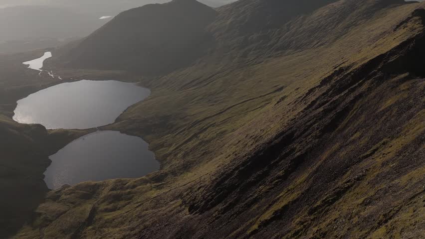 An expansive aerial view showcases a majestic mountain landscape. Two deep, still lakes are perfectly framed by the towering, ancient peaks under the gentle light of dawn.