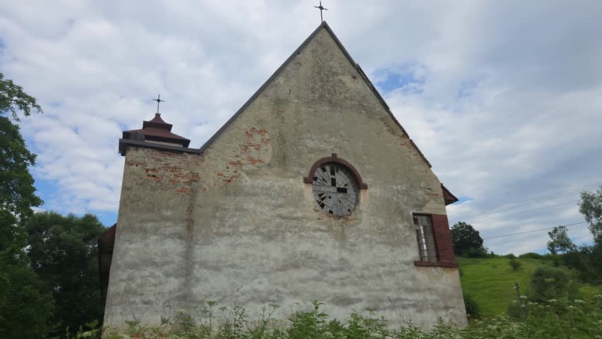 Abandoned brick church with a red roof and round window, standing in a lush green field surrounded by trees and a forest under a bright summer sky with white clouds