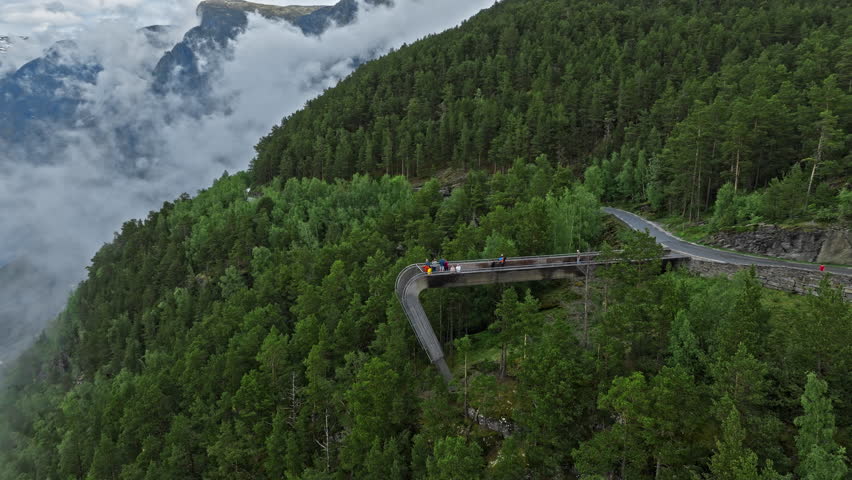 Tourists standing on the Stegastein viewpoint above Aurlandsfjord, Norway. Curved platform extends over forested cliffs with a mountain road, deep fjord, and low clouds creating a dramatic scenic travel view.
