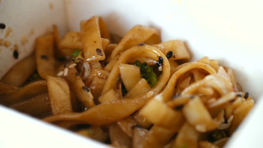 Close up chopsticks mixing hot asian wok noodles with broccoli, spring onions, sesame seeds inside white cardboard takeout box, representing quick, convenient lunch. Mixing takeout noodles in a box