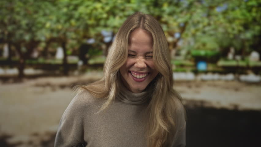 Woman blonde hair smiles showing teeth while standing on path in park under sunlight; happiness joy serenity relaxation.