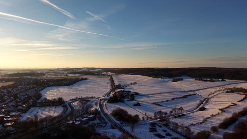 Snowy winter landscape with snow-covered fields at sunset time. Aerial forward wide shot. Roundabout surrounded by farm fields with white farmland fields.