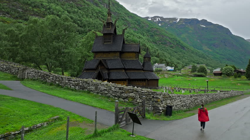 Tourist walking toward Borgund Stave Church on a quiet mountain day. Historic wooden church, bell tower, and stone walls sit in a green Norwegian valley under cloudy skies.