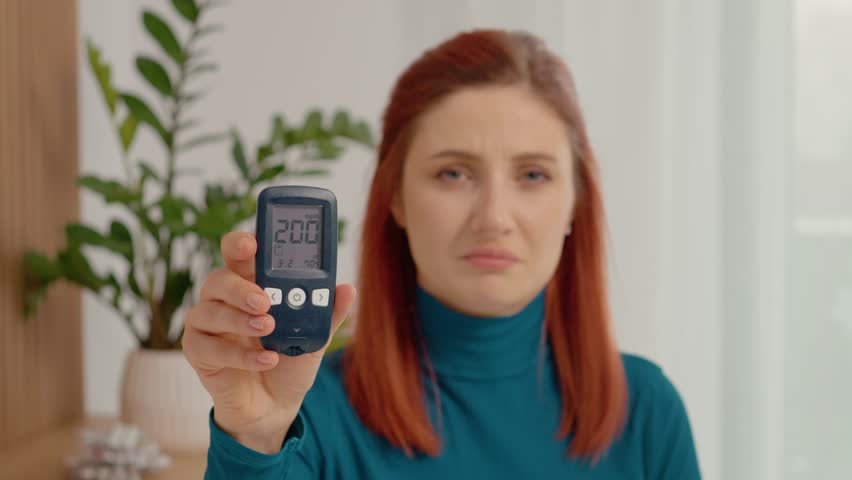 A woman looks at her glucometer reading of 200 mg dL with a shocked face. She is worried about her high blood sugar level in her home.