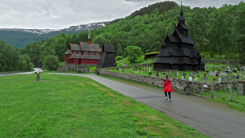 Tourist running toward Borgund Stave Church in Norway surrounded by green mountains. Woman visits historic wooden stave church and cemetery create a peaceful cultural travel scene on a cloudy day.