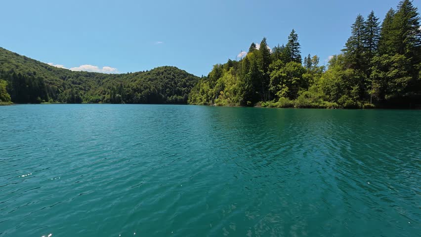 A wide view of the crystal-clear turquoise Kozjak Lake in Plitvice Lakes National Park. Lush green forested mountains surround the water under a bright blue summer sky.
