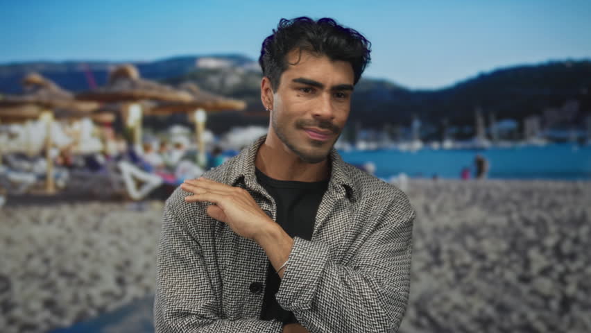Man gestures with hand pinching fingers near chin in front of a sunny beach outdoors; curiosity communication inquiry discussion.