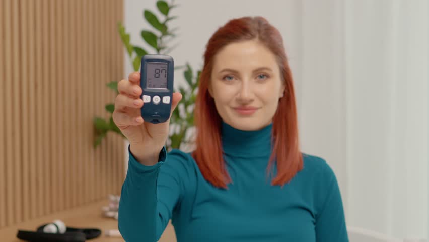A woman shares her glucometer reading of 87 mg dL and shows happiness in her living room. This shows positive diabetes management and monitoring of glucose levels.
