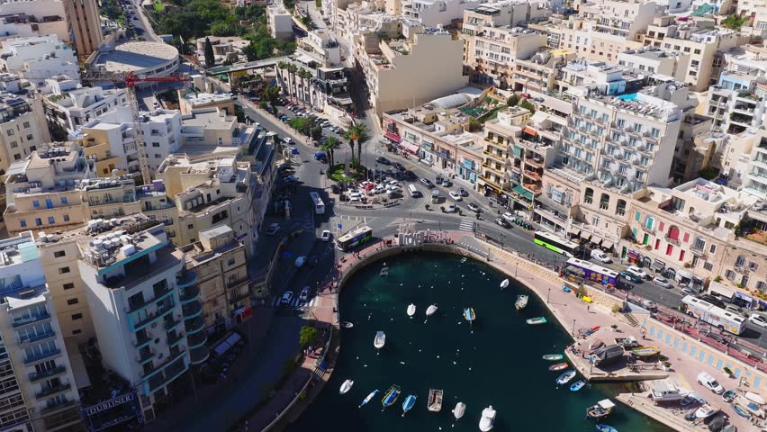 Aerial view of Sliema, Malta shows crescent harbor with boats, palm roundabout, promenade, limestone blocks, hotels, narrow streets, traffic, and turquoise water.