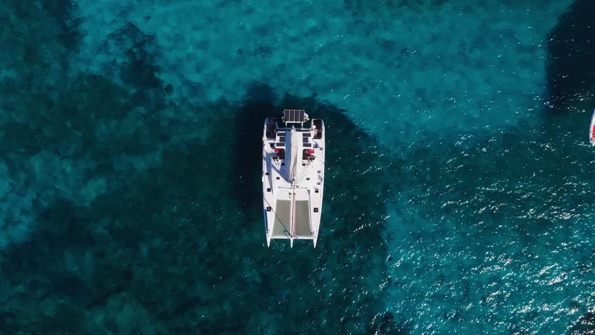 Aerial high angle view of a white catamaran anchored near Blue Lagoon, Comino, Malta. Twin hulls, nets, and solar panels show over seagrass and rocky seabed.