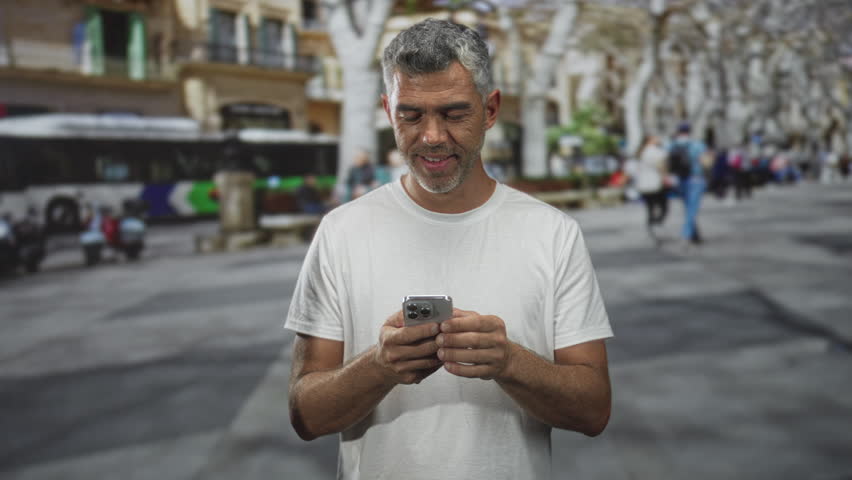 Man middle aged gray haired holding smartphone touching chin while looking up in a crowded urban street; quiet contemplation.