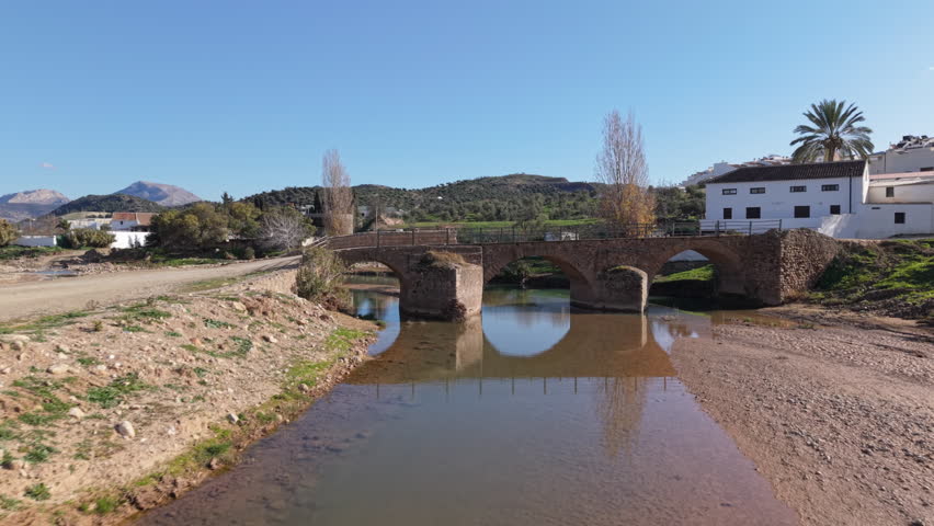 Aerial fly-in shot toward the ancient Roman bridge in Ardales, Spain. The drone approaches the well-preserved stone structure spanning a quiet river, surrounded by rolling hills and greenery. A serene and historic scene, ideal for cultural heritage, travel, or history-related content.

