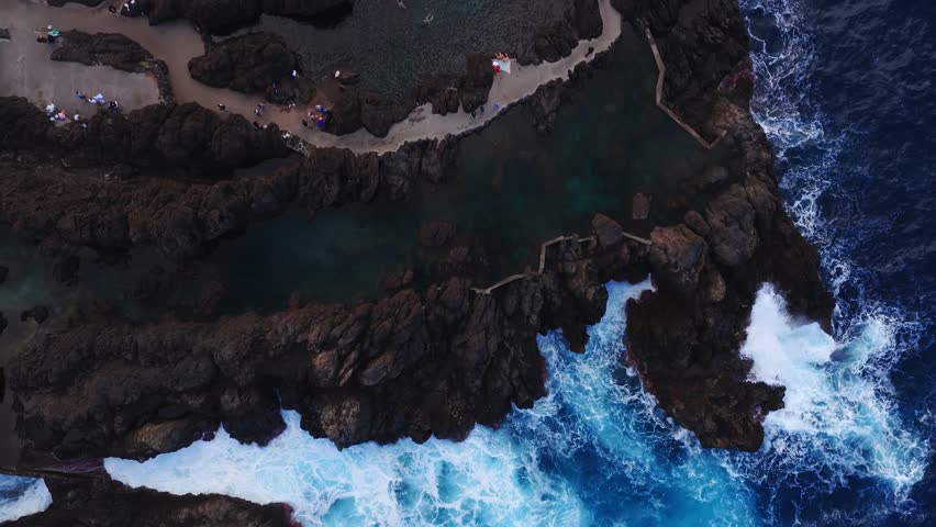Aerial top down view of Porto Moniz lava pools on Madeira, waves crash, swimmers float, visitors walk a paved path and circular platform in soft evening light.