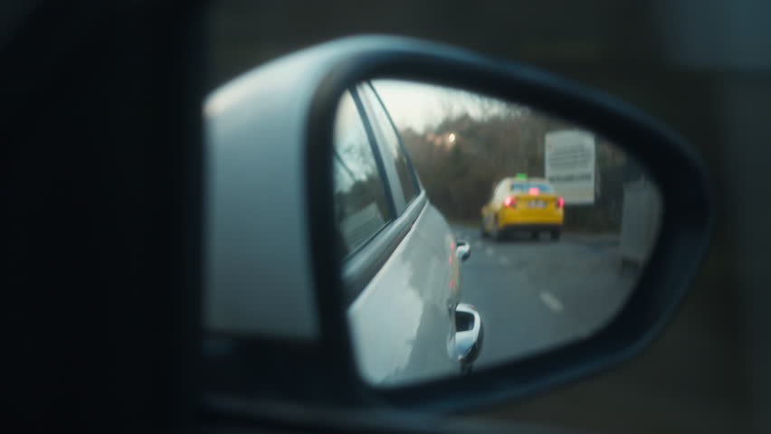 Rear side mirror view of a yellow car following on a curved road through a forested area, suggesting travel or pursuit.