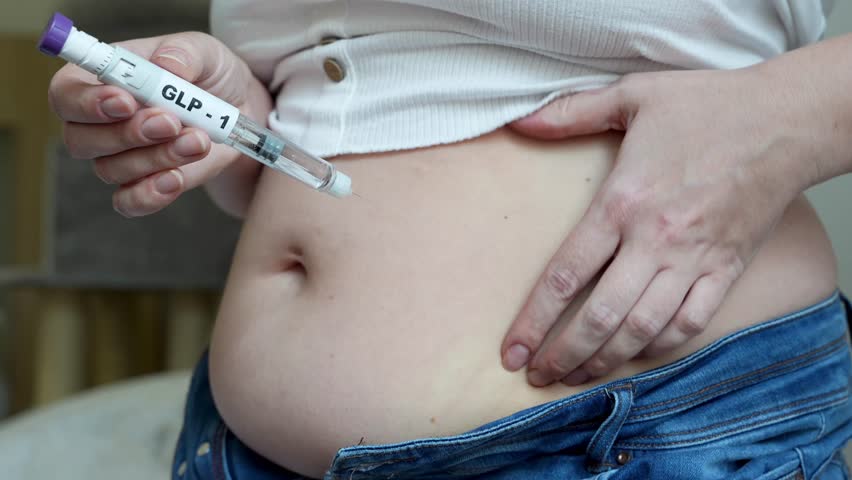 Woman prepares to administer GLP-1 medication via syringe into her abdomen, showcasing the self-injection process in a comfortable indoor environment