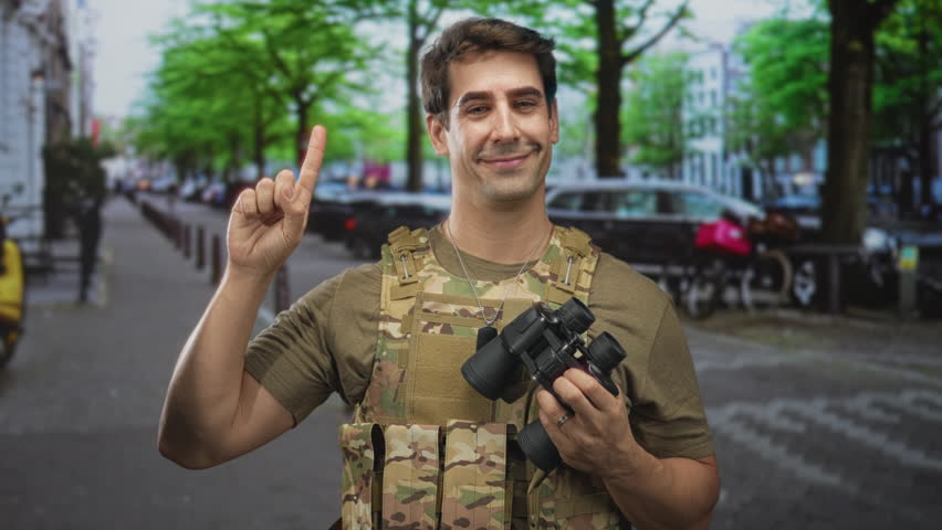 Man soldier wearing camouflage vest holding binoculars and pointing index finger on tree lined street with parked bikes; duty vigilance.