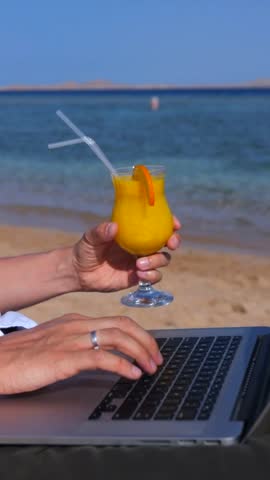 Male hands typing on a laptop keyboard while holding a glass of fresh orange juice on a tropical beach, illustrating the concept of a digital nomad lifestyle and remote work freedom