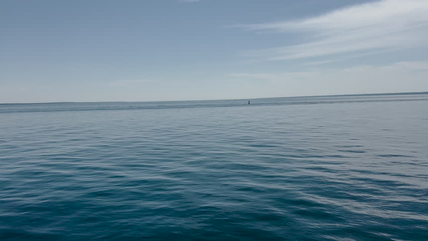 A steady, high-angle shot of gentle ripples on the surface of a deep blue lake with a clear horizon line under a soft summer sky. POV from a ferry boat on Lake Huron nearing Mackinaw Island, Michigan, USA.