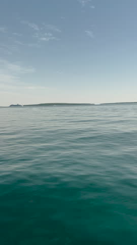 Vertical orientation of a steady, high-angle shot showing gentle ripples on the surface of a deep blue lake with a clear horizon line under a soft summer sky. POV from a ferry boat on Lake Huron approaching Mackinac Island, Michigan, USA.