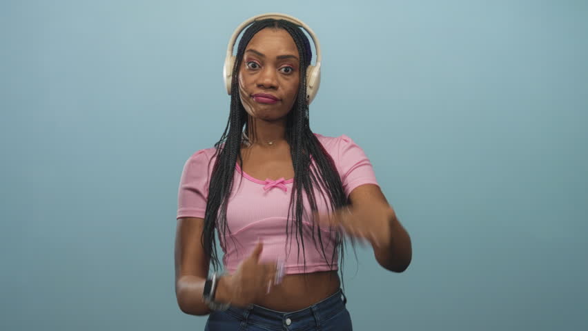 Woman with headphones wearing pink crop and shirt forms time out t with hands near chin in studio; indifference listening.