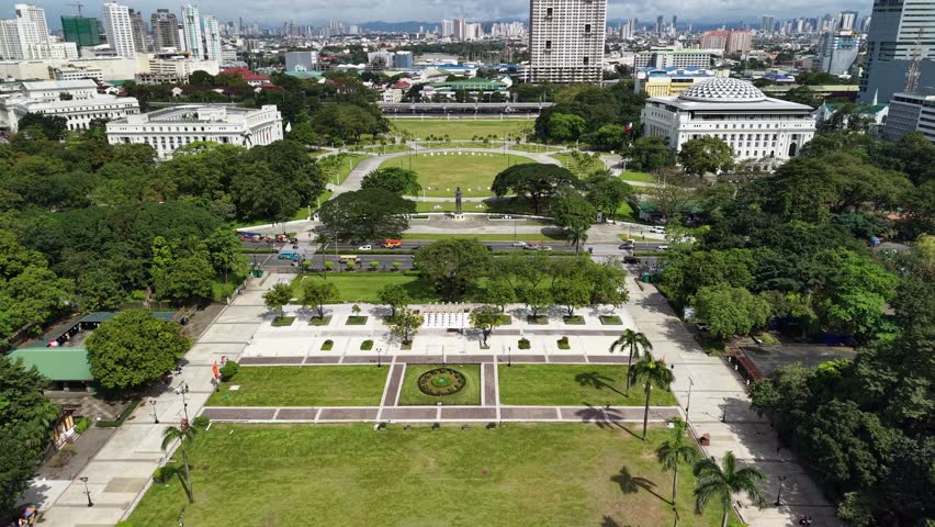 Stunning aerial perspective captures the iconic Rizal Park in Manila, Philippines, featuring vast manicured lawns and open plazas bordered by grand neoclassical architecture. The frame reveals the National Museum of Anthropology and Natural Museum buildings with their distinctive white facades and classical columns flanking both sides of the central green space. A prominent monument statue stands at the heart of the oval lawn, surrounded by walking paths and mature trees casting shadows across the grass. The foreground displays a spacious plaza with organized seating arrangements, while vehicles move along the busy road cutting through the scene. Lush tropical vegetation, including palm trees and dense canopies, punctuates the urban landscape. The midground showcases symmetrical pathways leading to central monuments, with pedestrians visible as small figures navigating the grounds. Beyond the immediate park area, the Manila cityscape rises dramatically with high-rise residential towers, commercial buildings, and modern skyscrapers creating a dense urban backdrop under partly cloudy skies. The lighting suggests daytime with natural illumination highlighting the contrast between green open spaces and surrounding urban development, creating a harmonious blend of historical heritage sites and contemporary metropolitan growth.