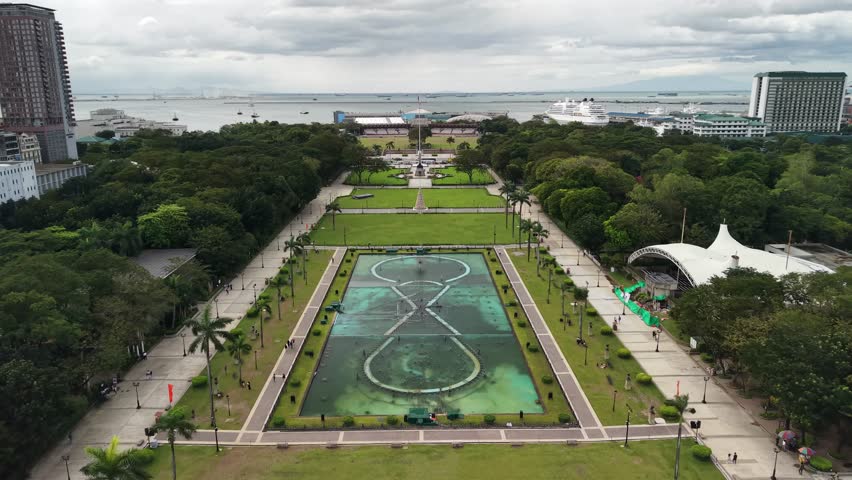 Cinematic aerial drone footage gliding forward over the iconic Rizal Park (Luneta) in Manila, Philippines, showcasing the stunning symmetrical layout of ornamental water features with vivid turquoise pools. The camera movement reveals geometric fountain designs - two large circular basins connected by an X-shaped water pattern creating mesmerizing reflections. Palm trees line both sides of the wide concrete walkways forming a perfectly balanced composition. Manicured green lawns stretch across the foreground and midground, dotted with visitors exploring the historic public space. A distinctive white tent-like pavilion structure stands on the right side among dense tropical foliage. Modern high-rise residential towers frame the left and right edges of the frame. The background opens to Manila Bay where multiple ships and vessels anchor in calm gray waters under a dramatic overcast sky with layered clouds. Dense tree canopy in rich green tones surrounds the formal garden layout. The Philippine flag pole rises in the distance beyond the fountains. Small groups of people stroll along pathways, creating a sense of scale and peaceful atmosphere in this celebrated national landmark and urban oasis.