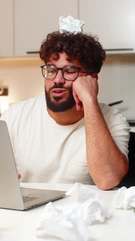 Arabian man at home writes ideas for project feels stuck looks at crumpled paper on desk breath slow. Young guy sitting at table frowns holds crumpled sheet on head critiques work plans next step