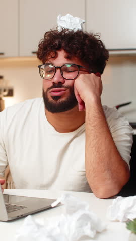 Arabian man at home writes ideas for project feels stuck looks at crumpled paper on desk breath slow. Young guy sitting at table frowns holds crumpled sheet on head critiques work plans next step