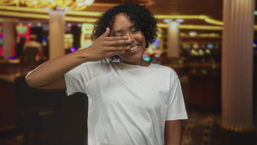 Woman covering eyes with hand and smiling behind fingers in casino building beside slot machines; excitement.