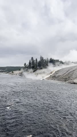 Static handheld video of the Firehole River flowing through Yellowstone, with textured current and ripples moving steadily downstream. Steam rises from the geothermal area along the riverbank to the right, drifting into the cool air and partially veiling the shoreline. Overcast skies and the contrast between dark water and pale steam create a moody hydrothermal landscape. No people or structures are visible.