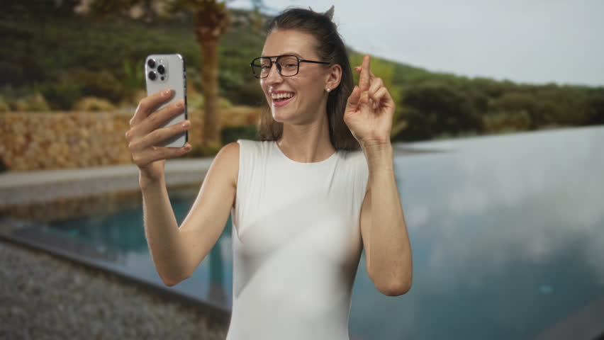 Woman holding smartphone and fingers crossed at building poolside, wearing white sleeveless top and glasses; optimism connection.