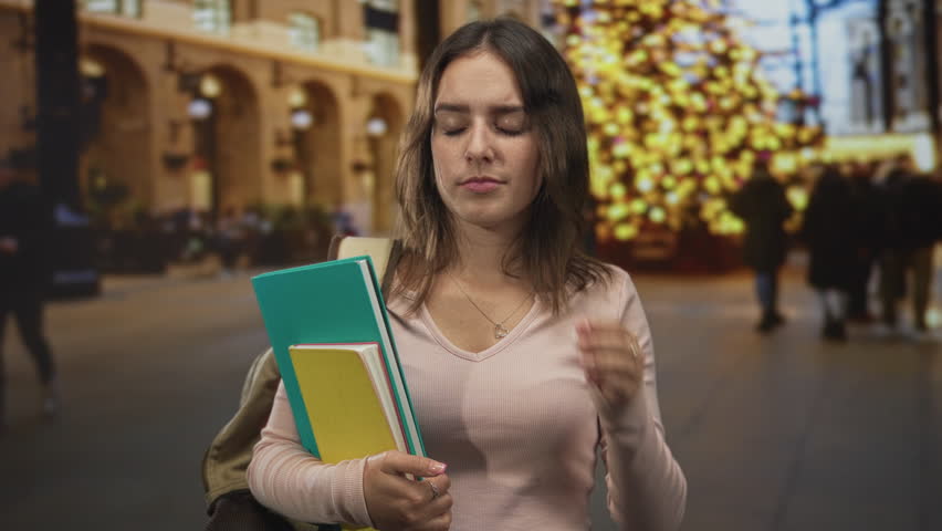 Woman holding notebooks and backpack, eyes closed, hand on temple on city street near holiday lights; stress tiredness.