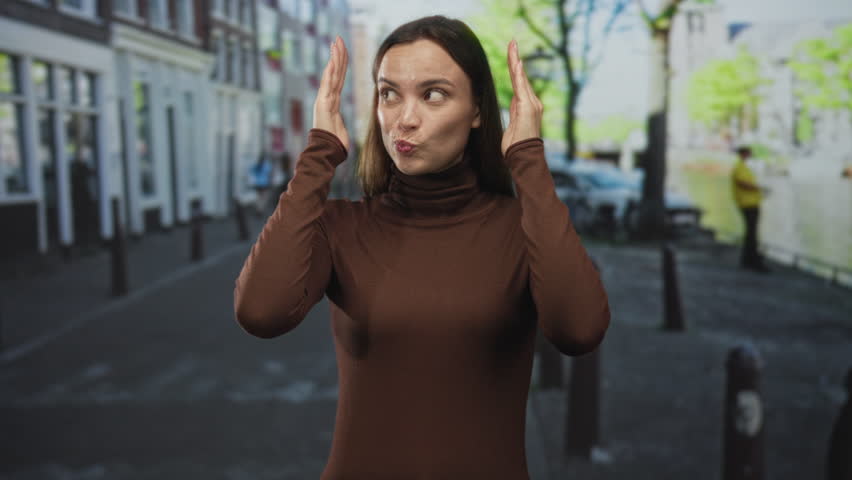 Young woman covering face with hands on city street by canal wearing brown turtleneck, alternating smile and frown gestures; surprise curiosity contemplation.