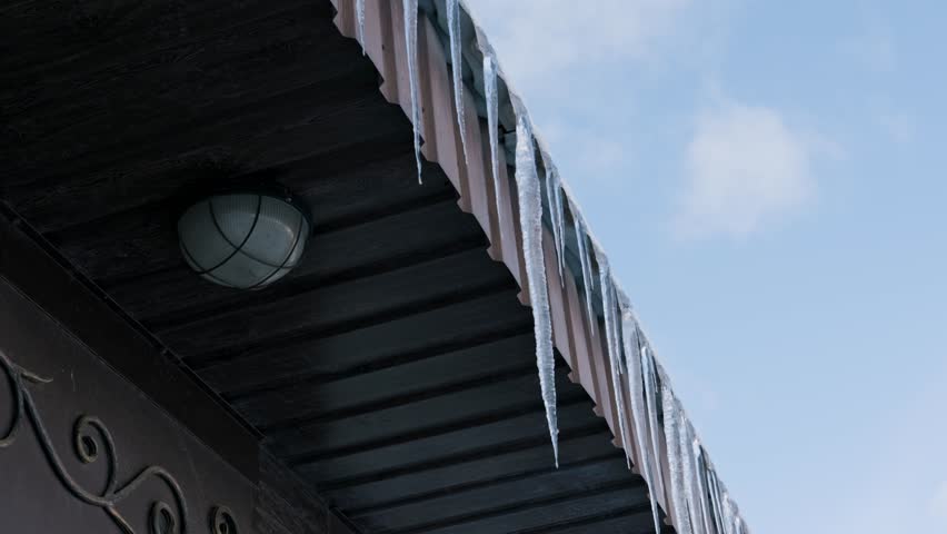 Long, sharp icicles hang in a row from the edge of a dark corrugated roof. A pale blue sky with soft white clouds is visible in the background, with a slow camera pan revealing the scene.