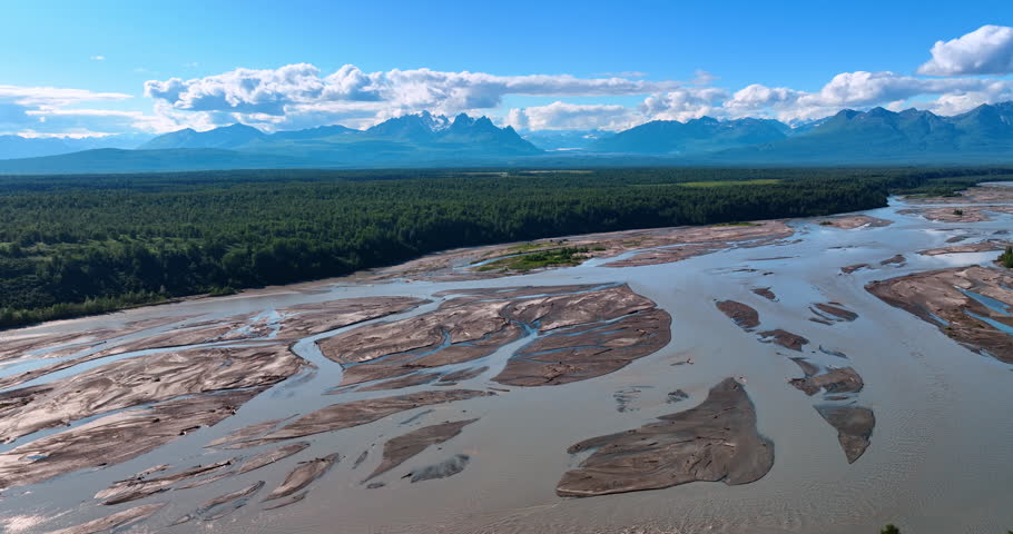 Stunning scenery of Alaska wilderness. Shallow river flows through the evergreen woods. Fluffy mountains hang over the mountain range at backdrop. Aerial view.